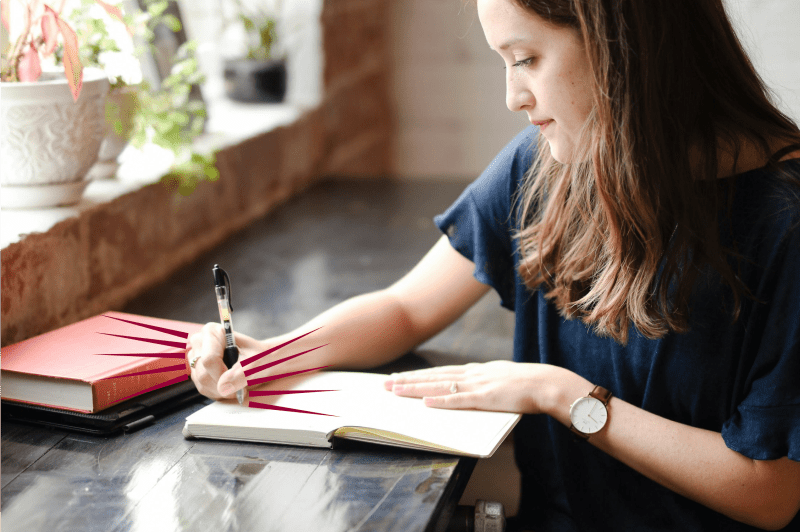 photo of a writer, sat at a black desk. in front of a windowsill filled with green plants